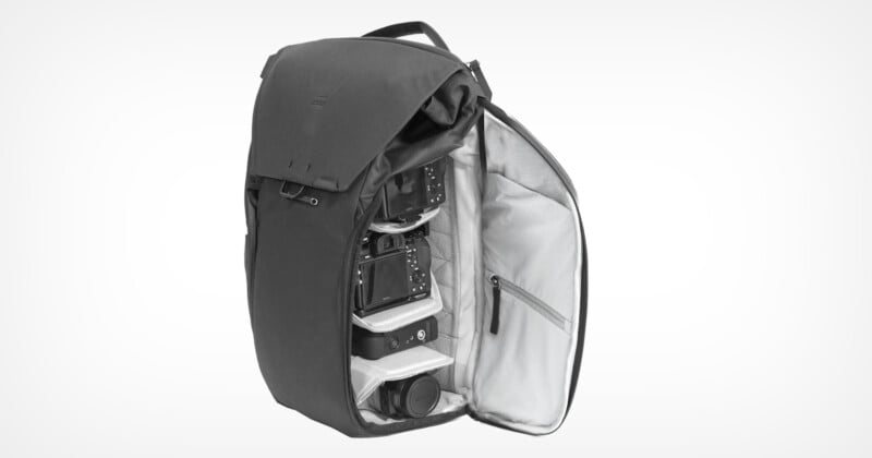 A black camera backpack opens to reveal organized compartments. Inside, camera bodies, lenses and accessories are neatly arranged. The interior is light gray, contrasting with the dark exterior. The backpack is located against a plain background.