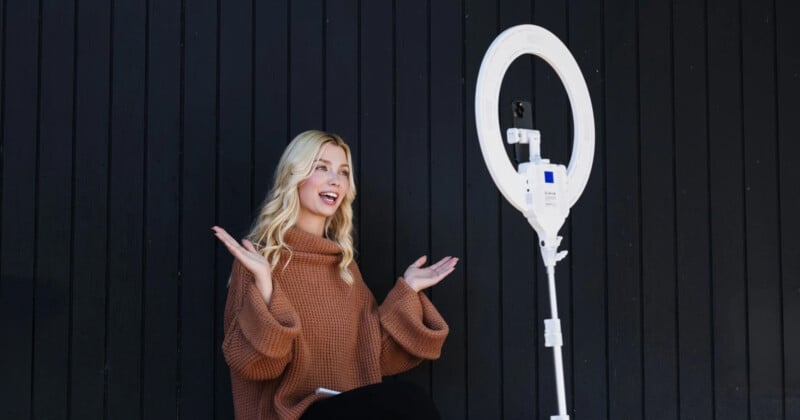 A man with long blonde hair and a brown sweater sits in front of a black wall and gestures at a smartphone on a ring light stand.