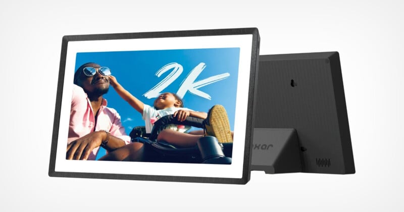 A digital photo frame shows an image of a person and a child smiling against a clear blue sky. The child playfully extends a hand toward the person. "2K" It is written in bold white letters on the picture. The frame has a simple, modern design.