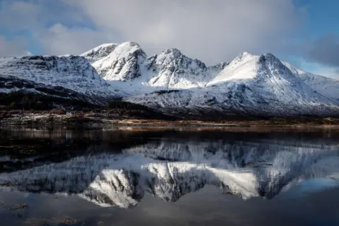 The peaks of the Lewis Love range are covered in snow, and are shown reflected in the waters of the loch.