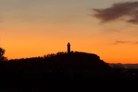 The Paul Weir Wallace Monument – ​​a tall, narrow structure – is silhouetted against the orange morning sky.