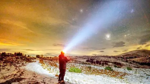 Alan O'Connor Man walks down a snowy mountain road in dark clothing with a head torch shining through the starry night sky.