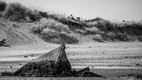 Edward Stephens black and white image of a beach with a dilapidated old boat sticking out of the sand and dunes in the background.