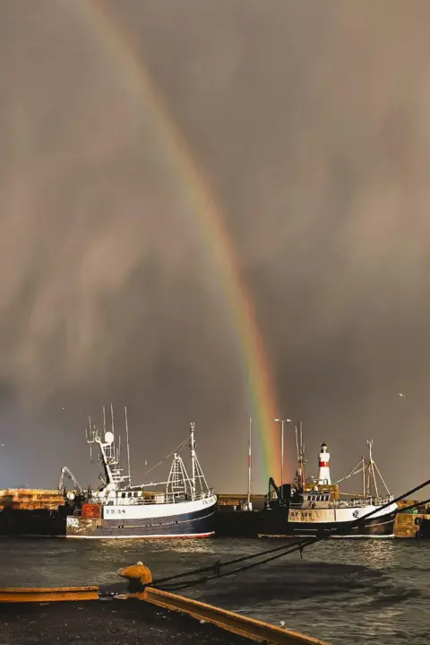 Two dark blue and white fishing trawlers in Ricci Harbor, Manila, with a rainbow arch and a red and white lighthouse behind them in a dark sky.
