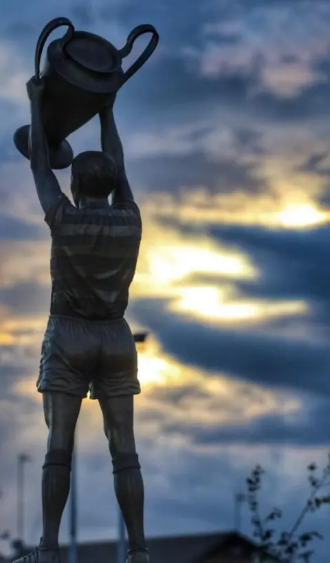 John Dyer statue of football player holding a large trophy with a blue and yellow sunset sky in the background.