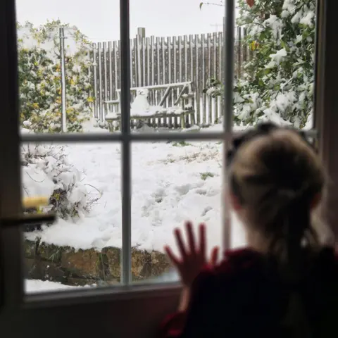Alex Watt sits outside in the snow and a little snowman looks out the window with one hand on the other.