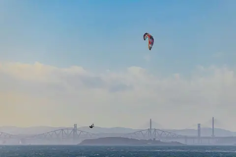 Keith Simpson kitesurfer mid-air with bright kite with bridges and blue sky and clouds in the distance.